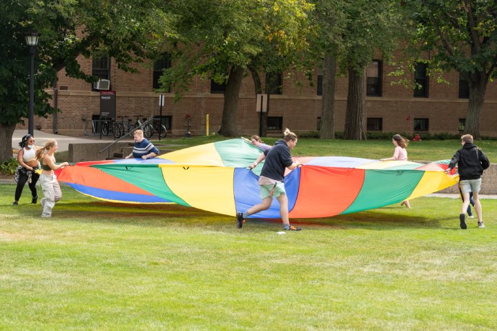A group of people playing with a large, colorful parachute on a grassy campus lawn. The participants hold the edges of the parachute and run around, with trees and a brick building in the background.